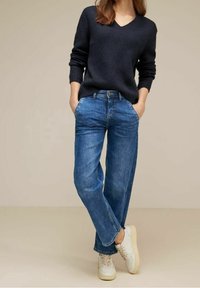 Pull en maille bleu marine avec un col en V, associé à un jean en denim bleu clair à coupe ample et des baskets blanches. Design simple, tissu texturé, style classique.