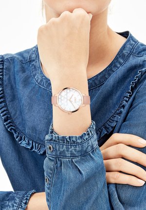 Roségoldene Uhr mit rundem Zifferblatt, irisierendem Ziffernblatt mit Herzmustern und einem weichen rosa Armband, getragen an einem jeansbedeckten Handgelenk.