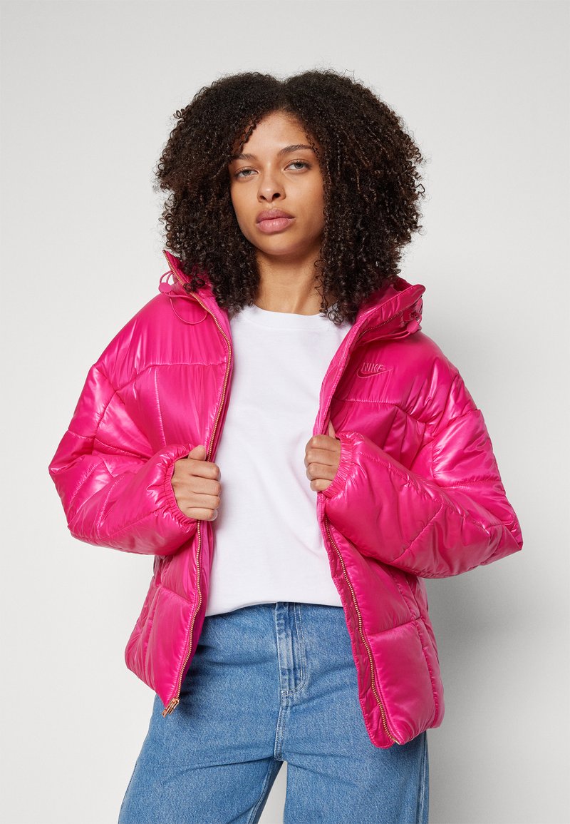 Puffige rosa Jacke mit glänzender Oberfläche, Kapuze und Reißverschluss. Mit elastischen Bündchen und Logo auf der Brust, getragen von einem Model in einem weißen Shirt.