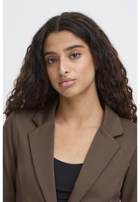 Brun kavaj med skräddarsydd design, texturerat tyg, urvinklade slag och ett svart underlager synligt. Modellen har långt, lockigt hår.