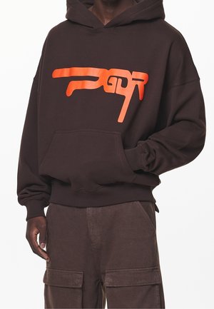 Dunkelbrauner Hoodie mit leuchtend orangefarbenem, stilisiertem Textlogo auf der Brust, kombiniert mit passenden dunkelbraunen Cargohosen.