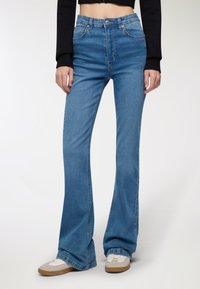 Spodnie dżinsowe z wysokim stanem i rozszerzonym dołem w jasnoniebieskim denimmie o gładkiej fakturze, z pięcioma kieszeniami i subtelnymi detalami stębnowania, zestawione z białymi sneakersami.