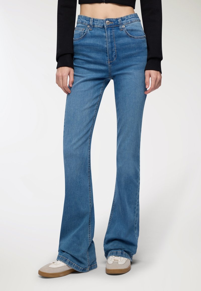 Spodnie dżinsowe z wysokim stanem i rozszerzonym dołem w jasnoniebieskim denimmie o gładkiej fakturze, z pięcioma kieszeniami i subtelnymi detalami stębnowania, zestawione z białymi sneakersami.