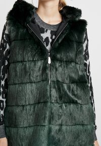 Grüner Kunstpelz-Weste mit Kapuze, ausgestattet mit einem Reißverschluss an der Vorderseite, einem gepanelten Design und einer glänzenden Textur. Getragen über einem gemusterten Pullover.