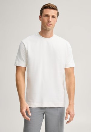 Homme debout devant un fond uni, portant un t-shirt blanc à manches courtes et un pantalon gris clair, regardant devant lui avec une expression neutre.