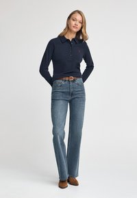 Navyblaues Langarm-Polohemd mit einem knöpfbaren Kragen und einem kleinen Logo, kombiniert mit hellblauen geraden Jeans und braunen Ankle-Boots.