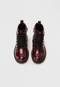 Bottes chelsea en cuir verni bordeaux avec des lacets noirs, des œillets dorés et une finition texturée. Semelle en caoutchouc noir durable avec une légère élévation.