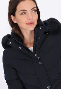 Marineblauwe pufferjack met een afneembare capuchon met bonttrim, rits- en drukknoopsluiting, en een gestikt patroon. Zachte, gladde stoftextuur zichtbaar.