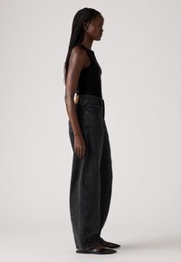 Un jean en denim noir taille haute avec une coupe ample, présentant une finition texturée et une patch en cuir beige à la taille. Associé à un top noir sans manches.