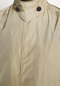 Beige lätt regnrock med hög krage, två svarta knappar och en texturerad yta, med synliga sömmar längs sömmarna.