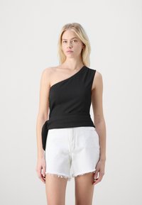 Svart ensidig topp med teksturert stoff og knyte-detajle på siden, sammen med hvite shorts med opprevet kant. Modellen står mot en nøytral bakgrunn.