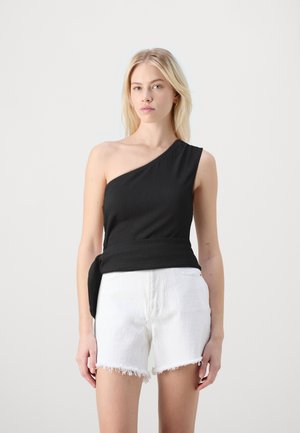 Juoda vieno peties viršutinis drabužis su tekstūrizuota medžiaga ir šoniniu raiščiu, derinamas su baltais šortais su nusidėvėjusia hemline. Modelis stovi prieš neutralų foną.