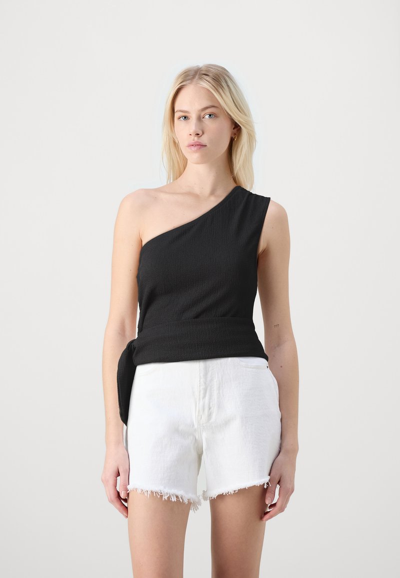 Svart ensidig topp med teksturert stoff og knyte-detajle på siden, sammen med hvite shorts med opprevet kant. Modellen står mot en nøytral bakgrunn.