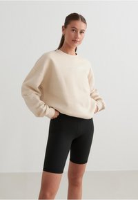 Beige mikina s kulatým výstřihem a jemným detailem loga, v kombinaci s černými cyklistickými šortkami. Směs bavlny, volný střih. Hladká textura.