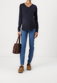 Kvinna som bär en marinblå V-ringad tröja med spetskant, blå jeans, bruna plattformsskor och håller en brun mocka bucket bag mot en vit bakgrund.