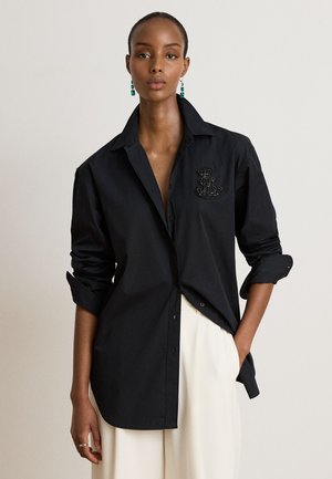 Femme portant une chemise noire boutonnée avec manches retroussées et un pantalon blanc, debout avec une main dans la poche devant un fond uni.