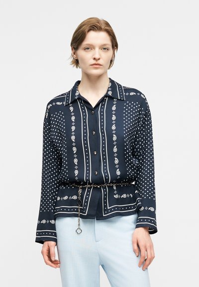 Femme aux cheveux courts portant une chemise boutonnée à motif bleu marine rentrée dans un pantalon bleu clair, avec une ceinture chaîne et une expression neutre.