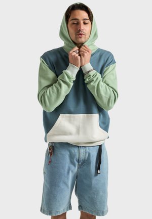 Junger Mann trägt einen blau-grünen Kapuzenpullover und Jeansshorts, zieht mit beiden Händen an den Kapuzenstrings vor einem hellen Hintergrund.