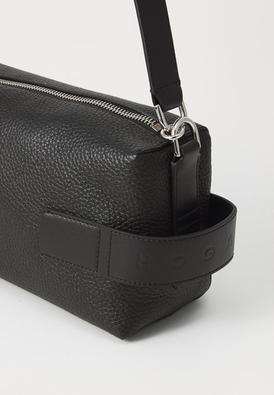 Geantă de umăr din piele neagră cu suprafață texturată, fermoar argintiu și inițiale ale brandului embosate pe o curea contrastantă. Formă rectangulară.