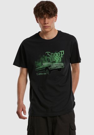 Jeune homme portant un T-shirt noir avec une voiture vintage verte et le texte « Snoop Dogg California » sur le devant, debout devant un fond uni.