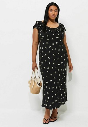 Frau trägt ein schwarzes, geblümtes Midikleid mit Rüschenärmel, hält eine beige geflochtene Handtasche und trägt schwarze Sandalen.