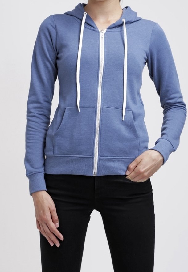 Personne portant un sweat à capuche bleu zippé avec des cordons blancs et un pantalon noir, debout avec une main dans la poche et l'autre reposant sur le côté.
