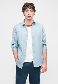 Ανοιχτό μπλε πουκάμισο με κουμπιά και μανίκια γυρισμένα, σε συνδυασμό με λευκό και πορτοκαλί μπλουζάκι με στρογγυλή λαιμόκοψη και σκούρο μπλε παντελόνι.