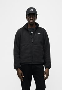 Άνδρας που φοράει μαύρο fleece μπουφάν, μαύρο T-shirt και μαύρο καπέλο, όλα με το λογότυπο The North Face, στέκεται μπροστά σε απλό λευκό φόντο.