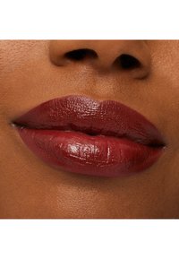 Tiefroter Lippenstift auf vollen Lippen aufgetragen, der eine glatte, glänzende Textur auf einem mitteltönigen braunen Hautton präsentiert. Dezente Lippenlinien sind sichtbar.