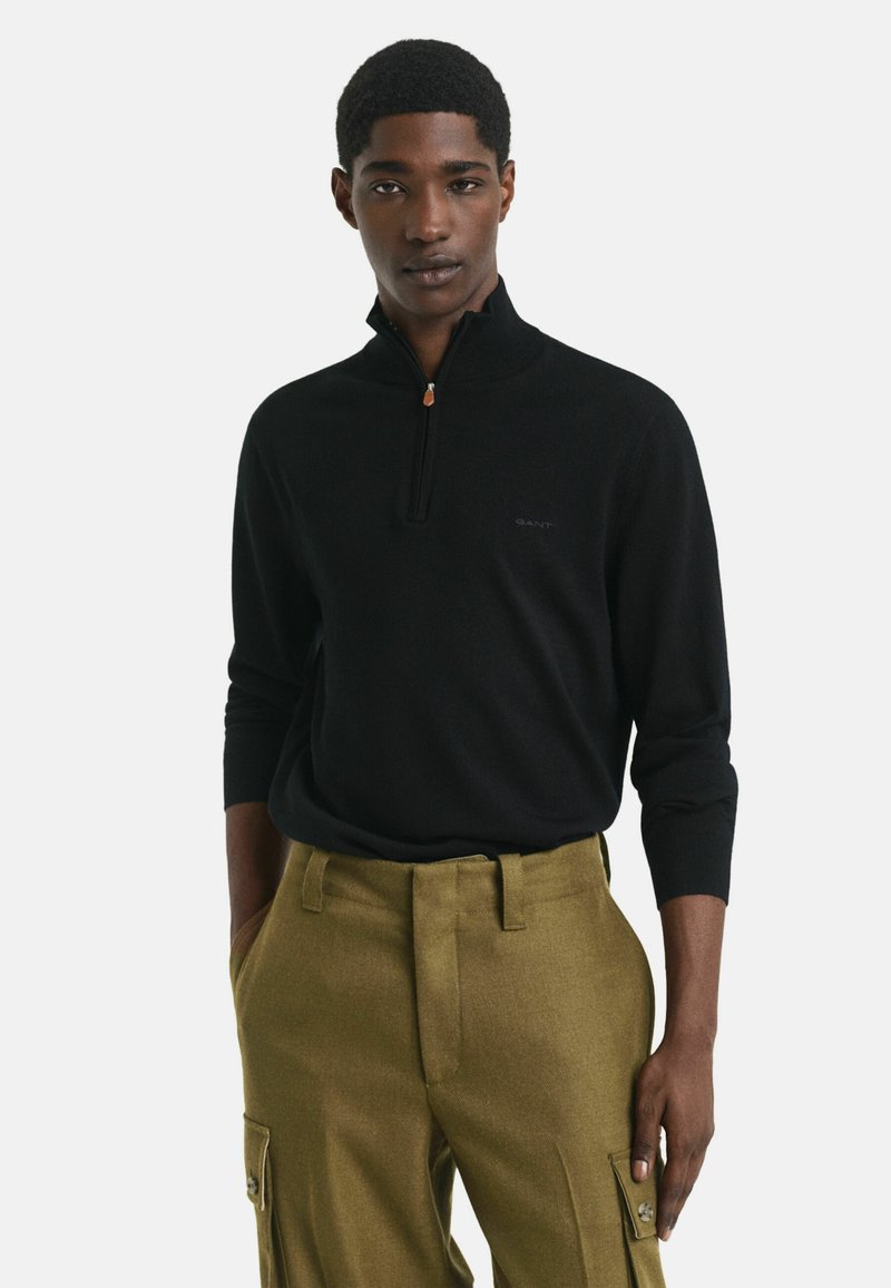 Homme portant un pull à col zippé noir à manches longues et un pantalon cargo vert olive, se tenant devant un fond clair uni.
