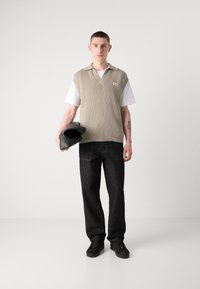 Bež pleteni jopič nad belo srajco, temno denim hlače, drži črn clutch, kratki lasje, nevtralna ozadja. Preproste oblikovalske značilnosti.