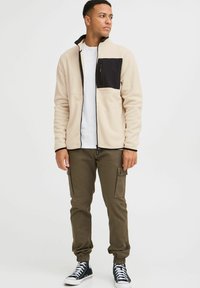 Chaqueta de forro polar en beige con un bolsillo en el pecho con cremallera negra. Combinada con pantalones cargo en verde oliva y zapatillas altas negras.