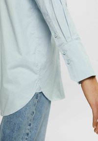 Ljusblå skjorta med knappar och långa ärmar, med manschetter som har två knappar och en rundad nederkant. Slät textur. Bärs över jeans.