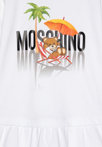 Camiseta blanca con un gráfico de un oso de peluche reclinado bajo una sombrilla naranja, con una palmera y el texto "Moschino" en negrita negra.