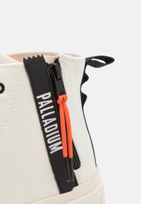 Witte hoge sneakers met een zwarte zijkant rits en een oranje trekkoord. Bevat een gestructureerde canvas bovenkant en een rubberen zool met het merkje.