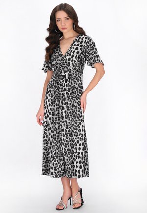 Frau mit langem welligem Haar, trägt Creolen, ein schwarz-weißes Leo-Print-Midikleid mit V-Ausschnitt und schwarze Sandaletten mit silbernen Riemen.