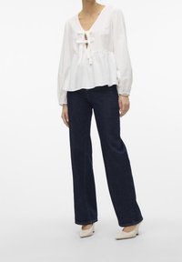 Blusa bianca a maniche lunghe con scollo a V e dettagli in chiffon, abbinata a jeans a zampa d'elefante blu scuro e scarpe con tacco crema.