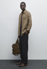 Khakifarbenes karierter Leinenhemd mit Knopfleiste, schwarzes T-Shirt darunter, lockere schwarze Hose, braune Wildledertasche, braune Flip-Flops, minimalistischer Hintergrund.