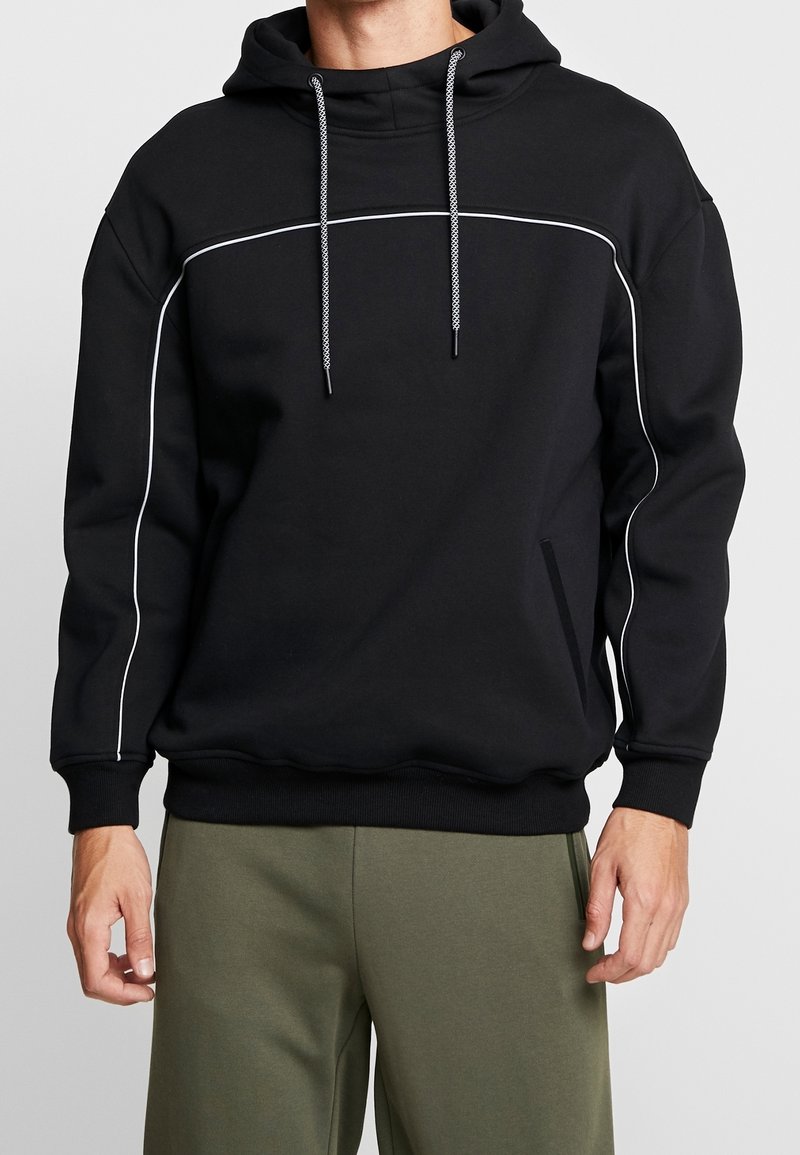Homme portant un sweat à capuche noir avec des passepoils blancs et un pantalon vert, debout devant un fond clair uni.