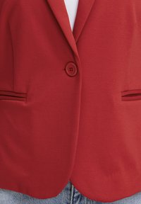Veste rouge boutonnée à la taille avec des revers en encoche, deux poches fendues horizontales, portée sur une chemise blanche et un jean bleu.