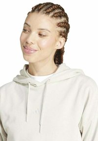 Giovane donna con capelli intrecciati che sorride, indossando un hoodie beige chiaro con lacci su uno sfondo bianco.