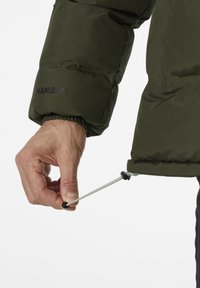 Groene pufferjack met elastische manchetten. Een hand trekt een trekkoord met een kleine zwarte stopper aan de onderkant strakker voor een goede pasvorm.