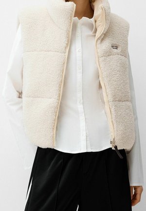 Femme portant une veste sherpa polaire couleur crème par-dessus une chemise blanche boutonnée et un pantalon noir, la veste étant à moitié dézippée.
