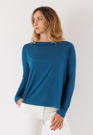 Maglia azzurra a maniche lunghe, tessuto morbido, vestibilità comoda, scollo a barca e maniche arrotolabili abbinata a pantaloni bianchi.