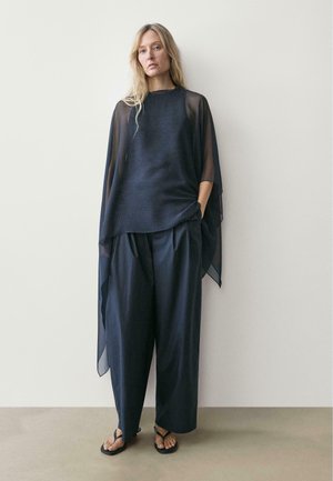 Kobieta o długich blond włosach, ubrana w luźną czarną przezroczystą bluzkę, szerokie czarne spodnie i czarne klapki, stojąca na tle jasnej, gładkiej ściany.