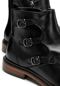 Botas de cuero negro hasta el tobillo con tres hebillas metálicas, puntera redonda y un acabado texturizado. Presentan un tacón de madera y costuras en contraste.
