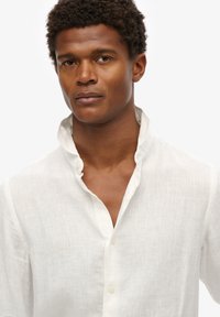 Camisa blanca de lino con cuello mao, con una textura sutil y una fila de botones, llevada por una persona con cabello corto y rizado.
