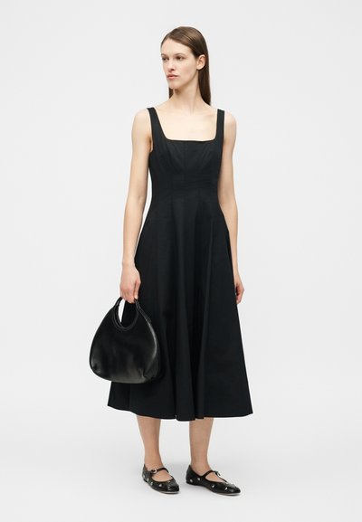 Γυναίκα που φοράει μαύρο μίντι φόρεμα χωρίς μανίκια, κρατώντας μαύρη δερμάτινη τσάντα και φοράει μαύρα επίπεδα παπούτσια με μεταλλικές λεπτομέρειες.