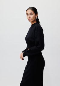 Svart kort genser med lange ermer, laget av et mykt stoff, sammen med et matchende tettsittende skjørt. Modellen står i profil mot en lys bakgrunn.