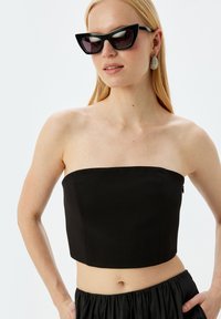 Musta olkaimeton crop top, jolla on tiukka muotoilu, sileä pinta ja suora kaula-aukko; yhdistettynä suuriin mustiin aurinkolaseihin.